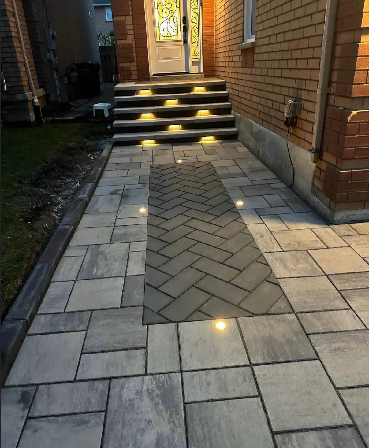 Night view of interlock walkway with backlit step lighting, herringbone runner accent, and in-ground LED pathway lights leading to red brick front door — D&D Exterior Finishing